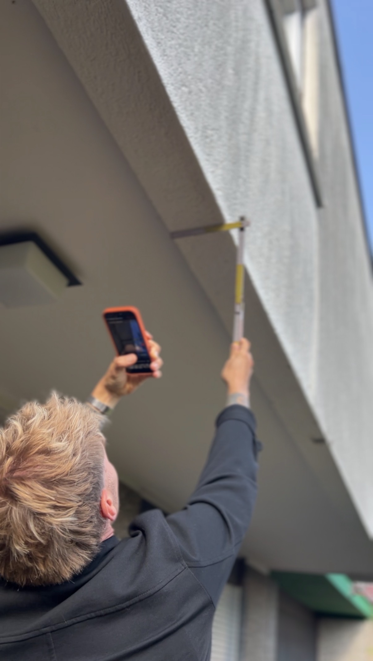 Gutachter misst bei der Begehung mit Zollstock und Smartphone ein Bauteil an der Fassade